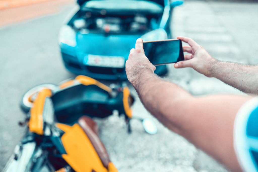 A man takes a photo on his smartphone of an accident scene between a car and a motorized scooter