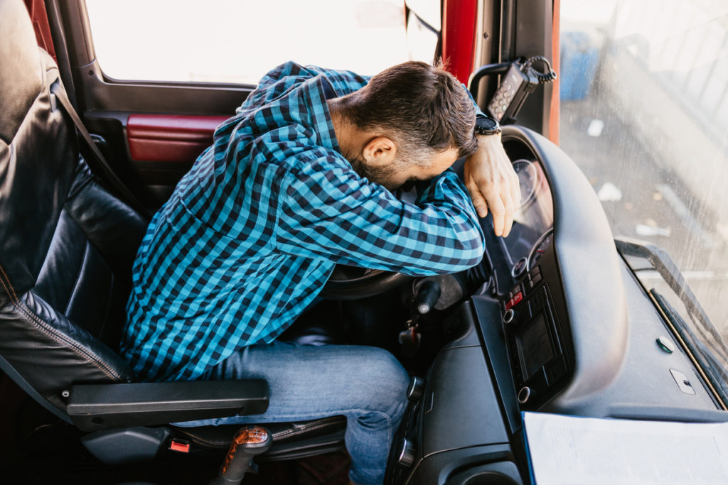 An exhausted male truck driver who is parked outside a warehouse, sleeps hunched over his stearing wheel