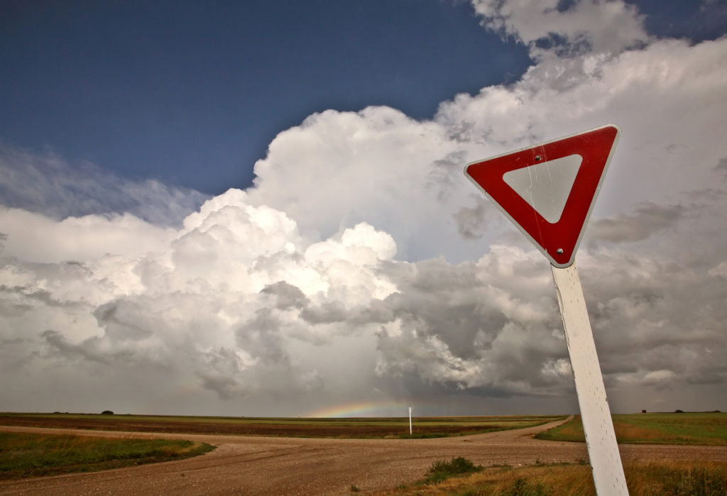 A traffic yield sign is shown on the side of a country crossroads with the sun off camera, but illuminating puffy clouds and a rainbow seen in the distance