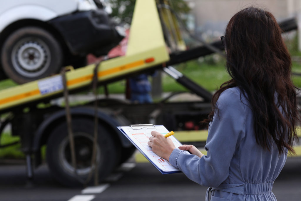 An adjuster making notes on a clipboard with a car being towed away in front of her