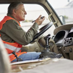 Side view of Hispanic man wearing casual clothing and reflective vest sitting in driver’s seat holding smart phone and listening.