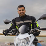 waist up shot of a man with latin features riding his motorcycle while is smiling and looking at the camera with a cloudy sky behind him