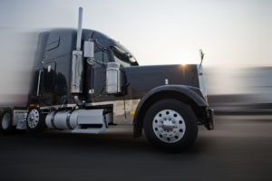 Profile of popular American dark bonnet classic big rig semi truck tractor with chrome and stainless steel accessories going on the road with sunshine in blurred background