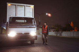 Full length view of early 30s Caucasian man in casual attire and reflective vest preparing to depart in delivery vehicle with headlights on at night.